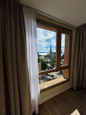 A room with a large window, curtains, and a view of a church and buildings. - Vista y cochera en pleno centro (San Carlos de Bariloche)