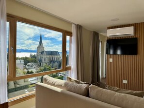 A modern living room with a large window, a flat-screen TV, and a wooden wall panel. - Vista y cochera en pleno centro (San Carlos de Bariloche)