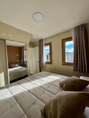 A bedroom with a bed, a mirror, a door, and two windows with curtains. - Vista y cochera en pleno centro (San Carlos de Bariloche)