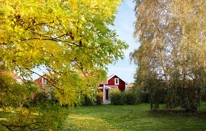 Une grange rouge avec une porte blanche, entourée d’une cour verdoyante et d’arbres aux feuilles jaunes.