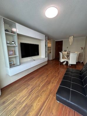 Ein modernes Wohnzimmer mit einem flachbildfernseher, der an einem weißen Schrank befestigt ist, einer schwarzen Ledersofa und einem Essbereich mit einer Kristallleuchter.