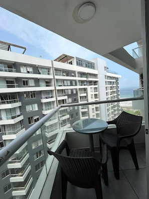 Ein Balkon mit einem Glastisch und zwei Korbstühlen, mit Blick auf ein Hochhaus und den Ozean.