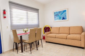 Een woonkamer met een beige bank, een eettafel met stoelen, een brandblusser en een lijst met een abstract schilderij.