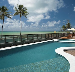A pool with a curved edge, surrounded by a railing, with palm trees and a beach view in the background. - Apartment in the luxurious boutique hotel Ritz Suites, fully equipped! (Maceió)