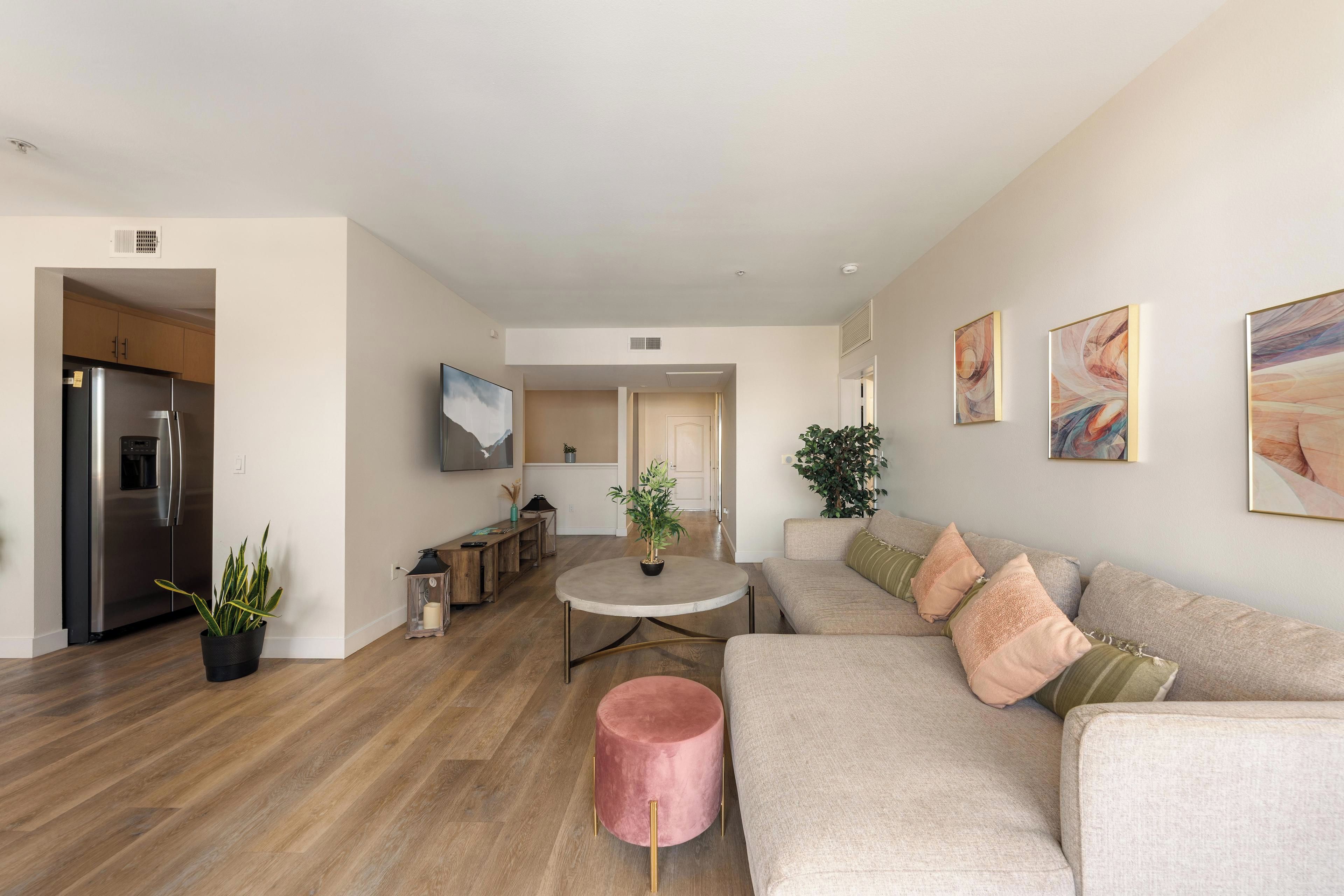 Ein modernes Wohnzimmer mit einem beigefarbenen Sofa, einem pinkfarbenen Hocker und einem runden Couchtisch. Es gibt eine Küchenzeile mit einem Kühlschrank und einem Esstisch.