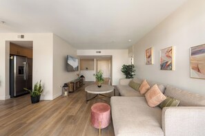 Ein modernes Wohnzimmer mit einem beigefarbenen Sofa, einem pinkfarbenen Hocker und einem runden Couchtisch. Es gibt eine Küchenzeile mit einem Kühlschrank und einem Esstisch.