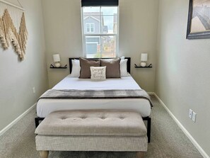 Une chambre à coucher avec un lit, des tables de chevet, une fenêtre et une décoration murale en macramé.