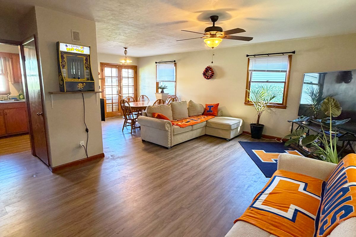 A living room with a sofa, a coffee table, and a ceiling fan. There is a dining area with a table and chairs.