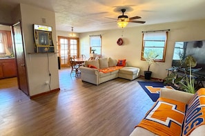 A living room with a sofa, a coffee table, and a ceiling fan. There is a dining area with a table and chairs. - The Illini House - UIUC Game Day Getaway (Urbana)