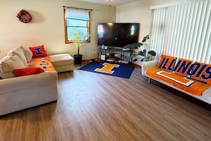 A living room with a sofa, a television, and a plant. - The Illini House - UIUC Game Day Getaway (Urbana)