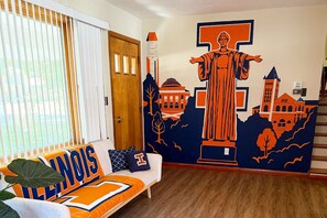 A room with a large mural, a sofa with a 'ILLINOIS' pillow, and a wooden door. - The Illini House - UIUC Game Day Getaway (Urbana)