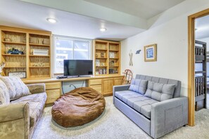 Ein Wohnzimmer mit Fernseher, zwei Sofas, einem braunen Hocker und Holzregalen mit diversen Gegenständen.
