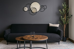 Un salon moderne avec un canapé gris foncé, deux petites tables rondes en bois et un élément décoratif mural.