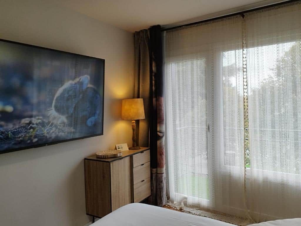 Ein Hotelzimmer mit Bett, Nachttisch mit Lampe, einem Bild an der Wand und einem Fenster mit durchsichtigen Vorhängen.