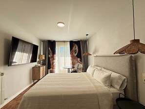 A hotel room with a bed, a television, a small table with a lamp, and a window with curtains.