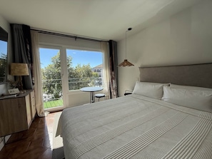 A bedroom with a large bed, a desk, a chair, and a small table. There is a balcony with a view of greenery and a building.