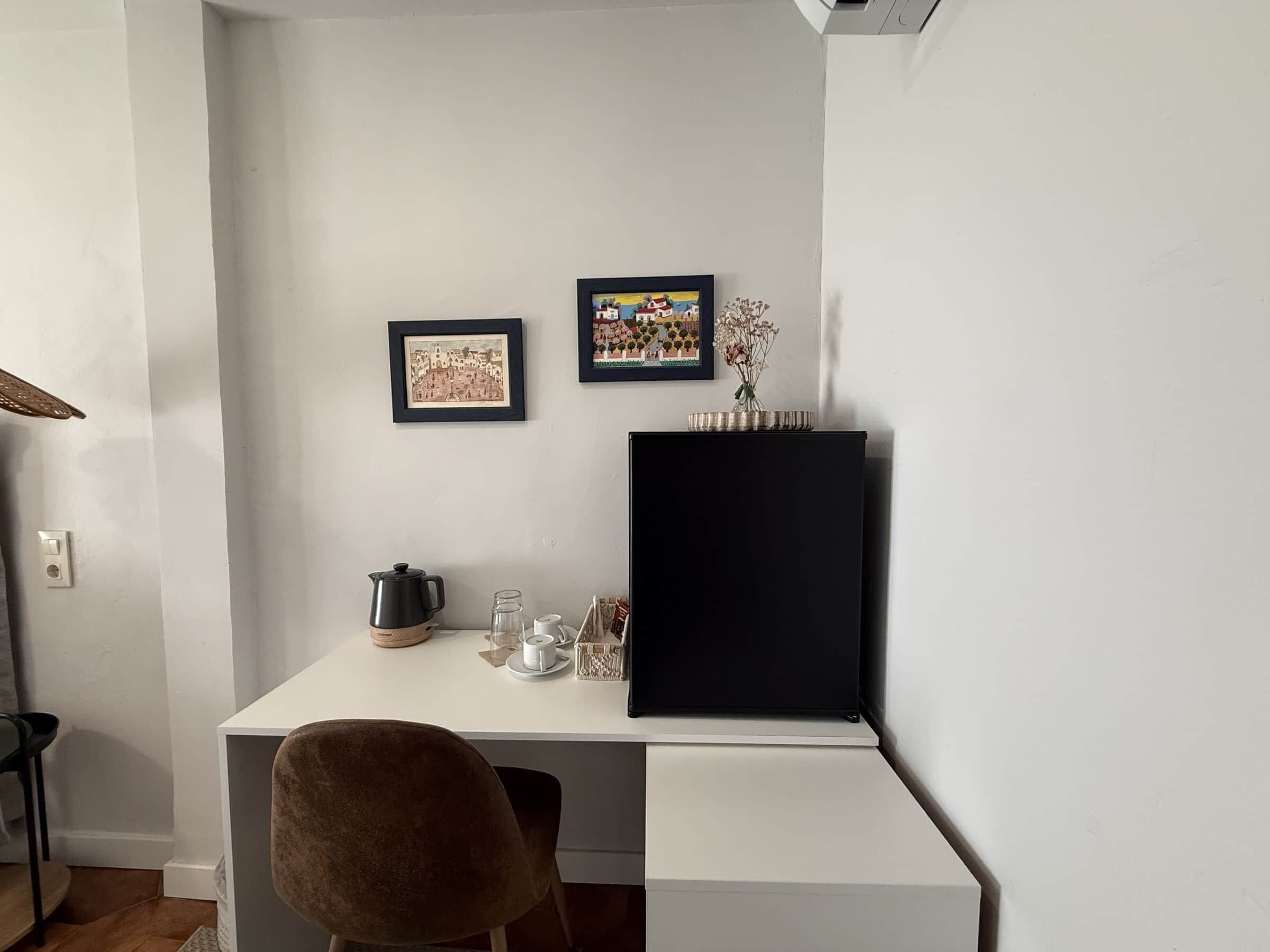 Un escritorio blanco con un monitor negro, dos cuadros enmarcados, un florero con flores y una cafetera.