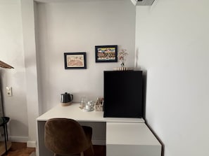 A white desk with a black monitor, two framed pictures, a vase with flowers, and a coffee pot.