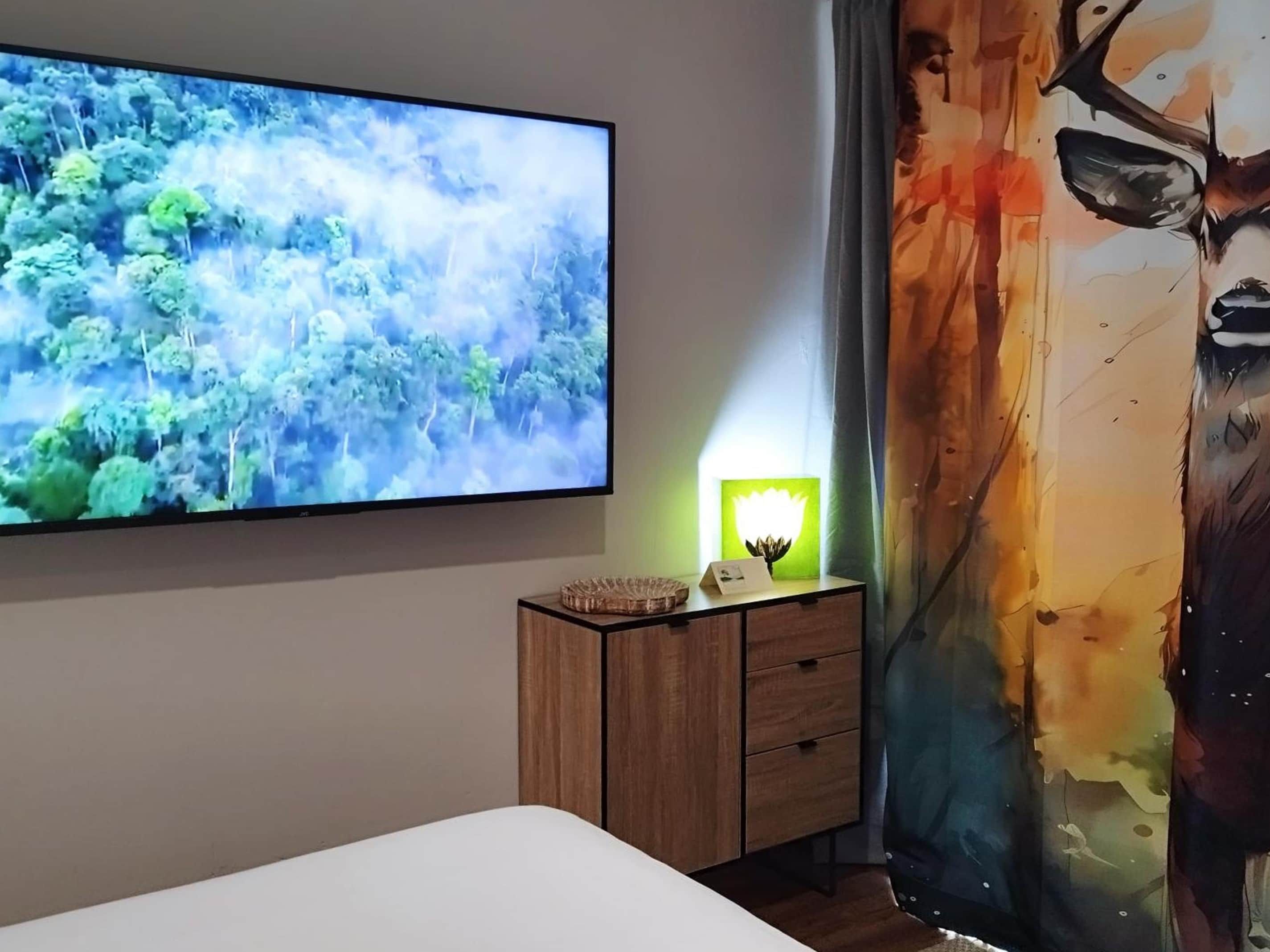 A bedroom with a large flat-screen TV mounted on the wall, a bedside table with a lamp, and a bed.