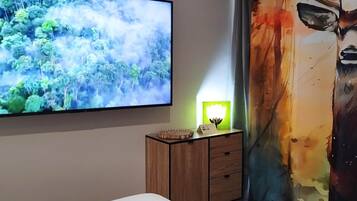A bedroom with a large flat-screen TV mounted on the wall, a bedside table with a lamp, and a bed.