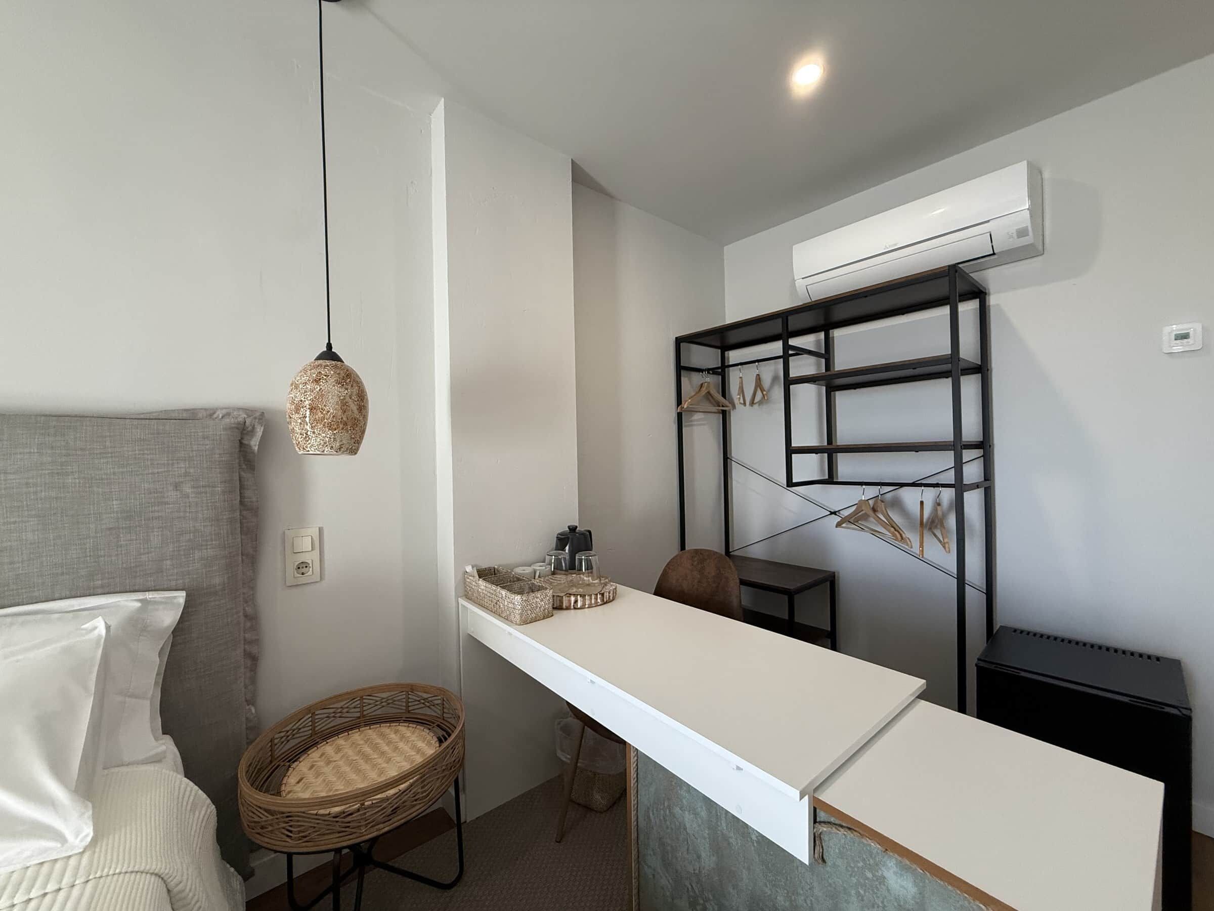A compact room with a bed, a white desk, a hanging light fixture, a wall-mounted air conditioner, and a black storage unit.
