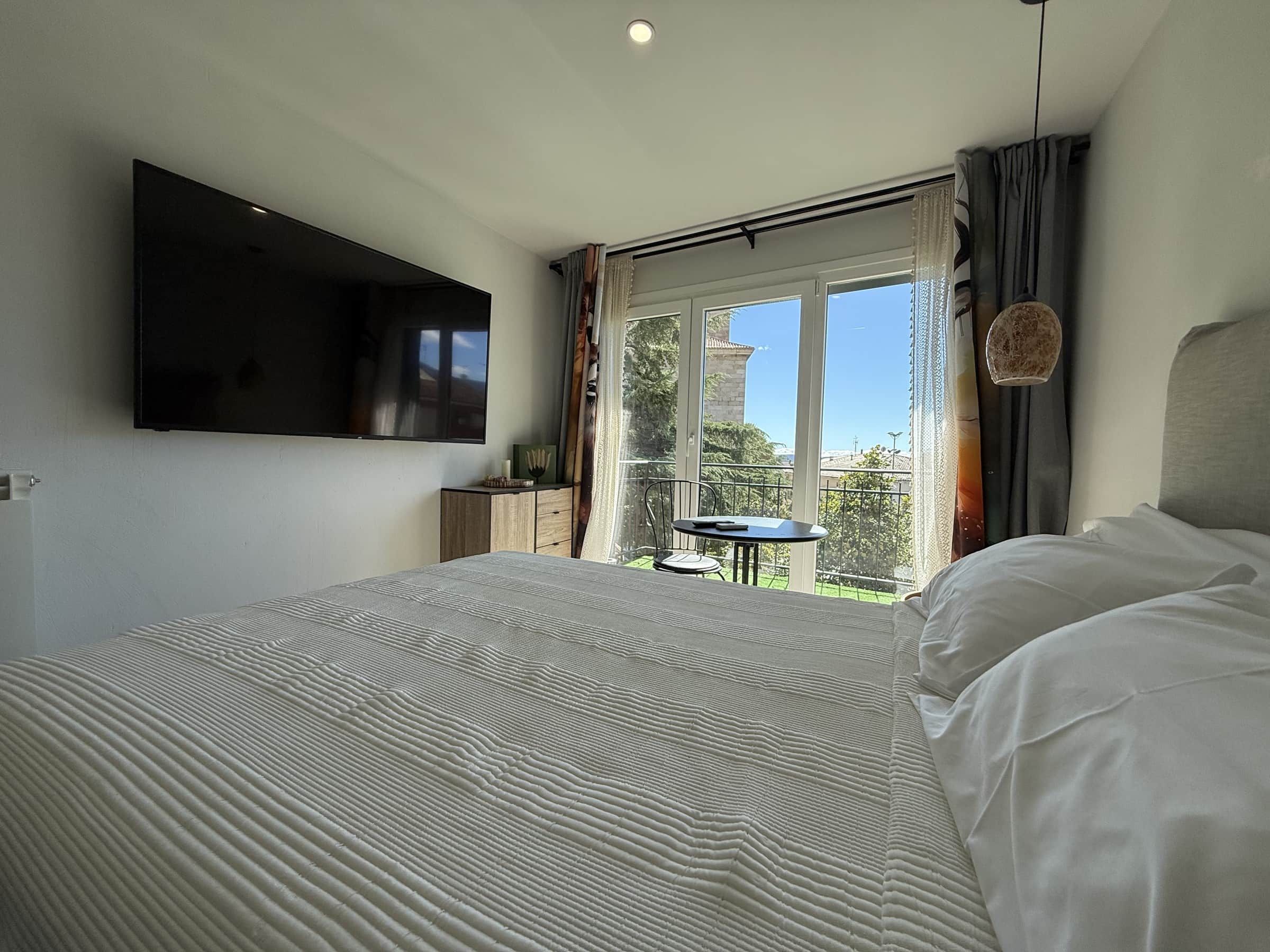 Un dormitorio con cama, televisor de pantalla plana en la pared, una mesita con silla y un balcón con vistas.