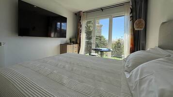 Un dormitorio con cama, televisor de pantalla plana en la pared, una mesita con silla y un balcón con vistas.