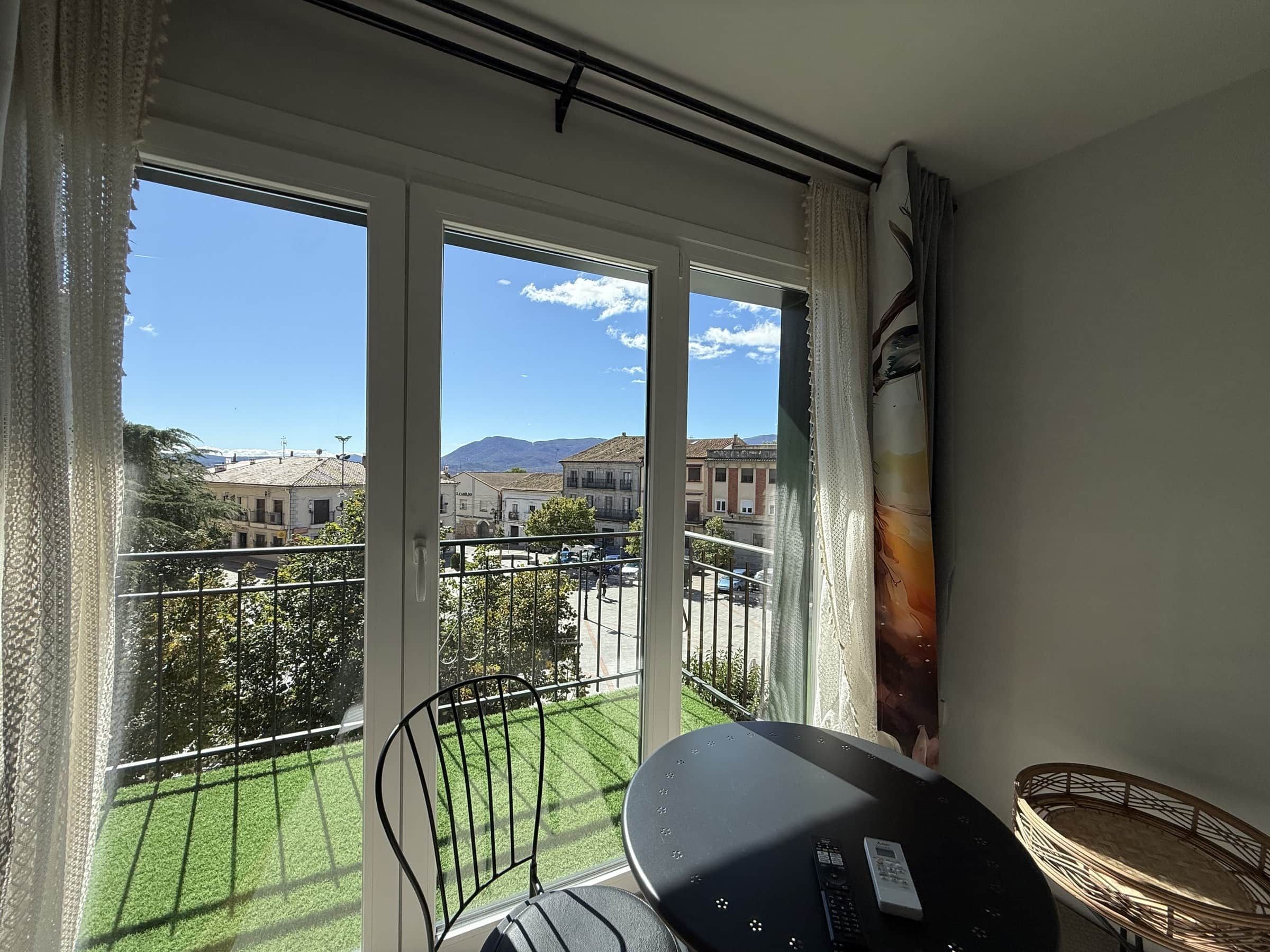 Un balcón con vista a un pueblo, una mesa con una silla y una ventana con cortinas.