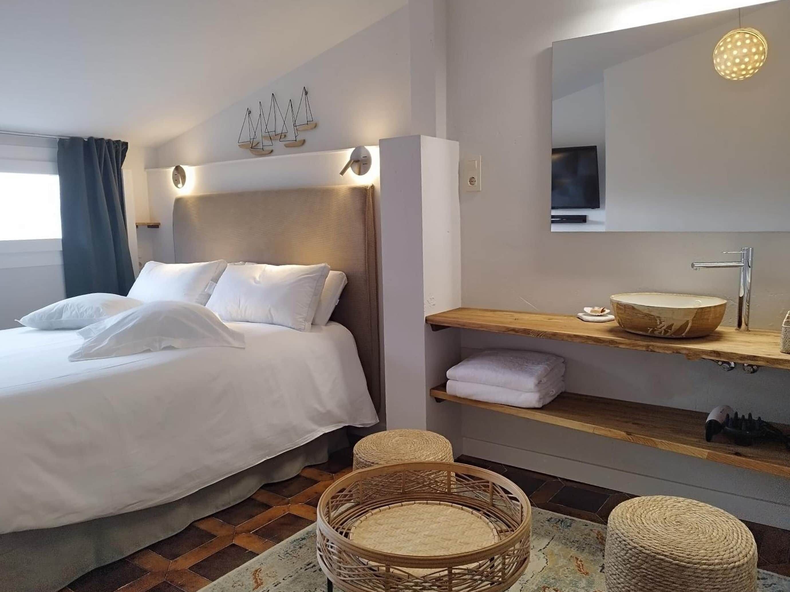 Una habitación de hotel con una cama, un estante de madera con un lavabo de madera redondo, un espejo y una lámpara de pared.