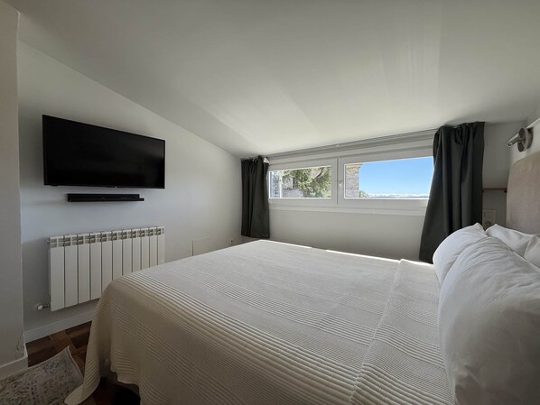 A bedroom with a bed, a television mounted on the wall, a window with curtains, and a radiator.
