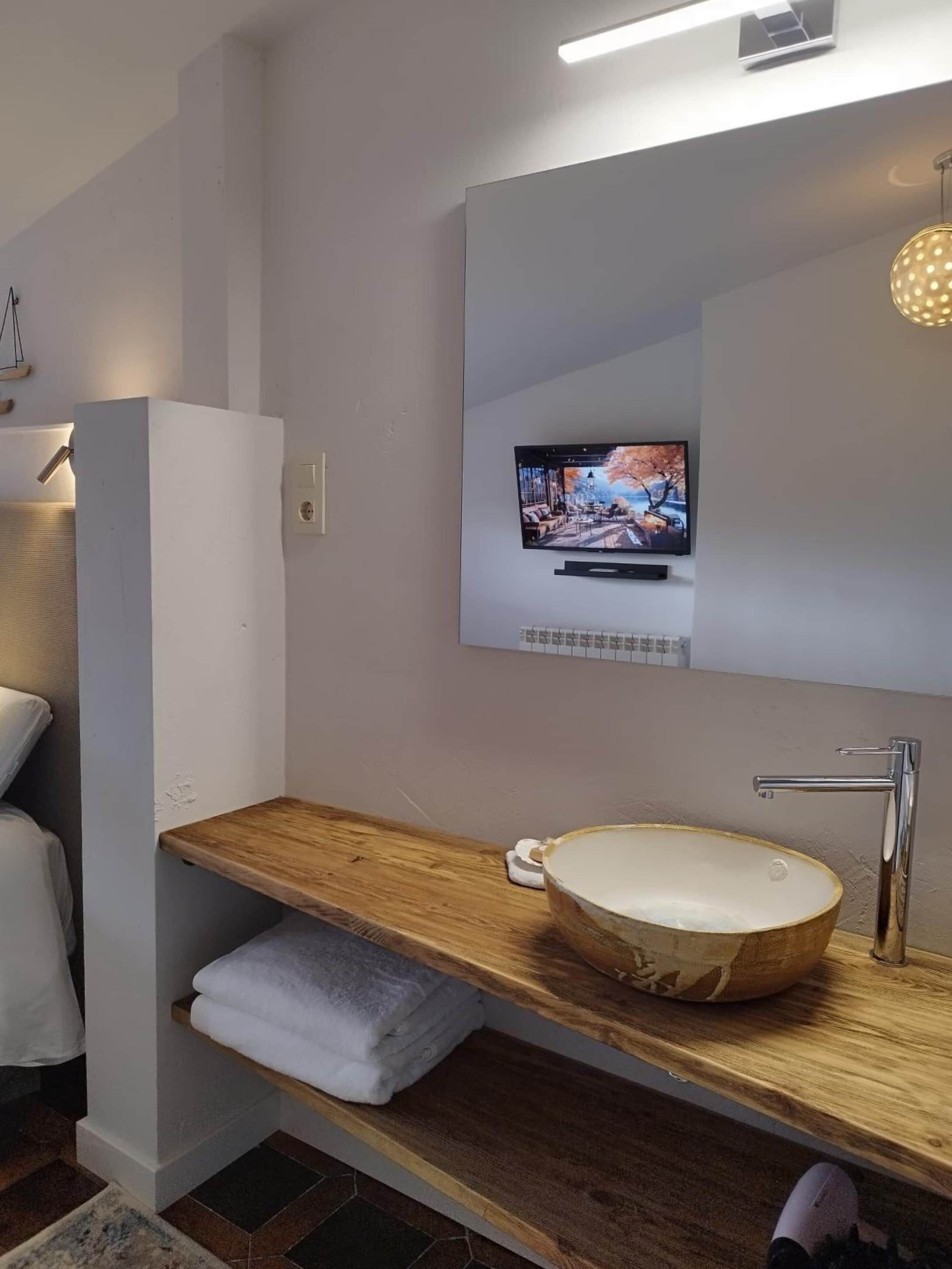 A modern bathroom with a wooden vanity, a large mirror, a television above the mirror, and a built-in sink.