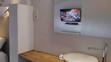 A modern bathroom with a wooden vanity, a large mirror, a television above the mirror, and a built-in sink.