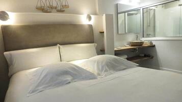 A bed with white linens, a headboard with built-in lighting, a bedside table with a mirror, and a wall-mounted shelf.