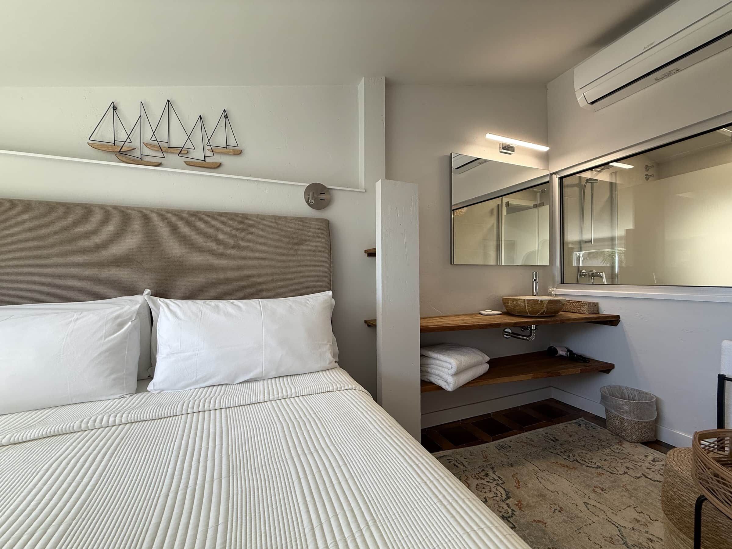 Une chambre d’hôtel avec un lit, une tête de lit en bois, un miroir fixé au mur, une étagère en bois avec des serviettes et une petite fenêtre.