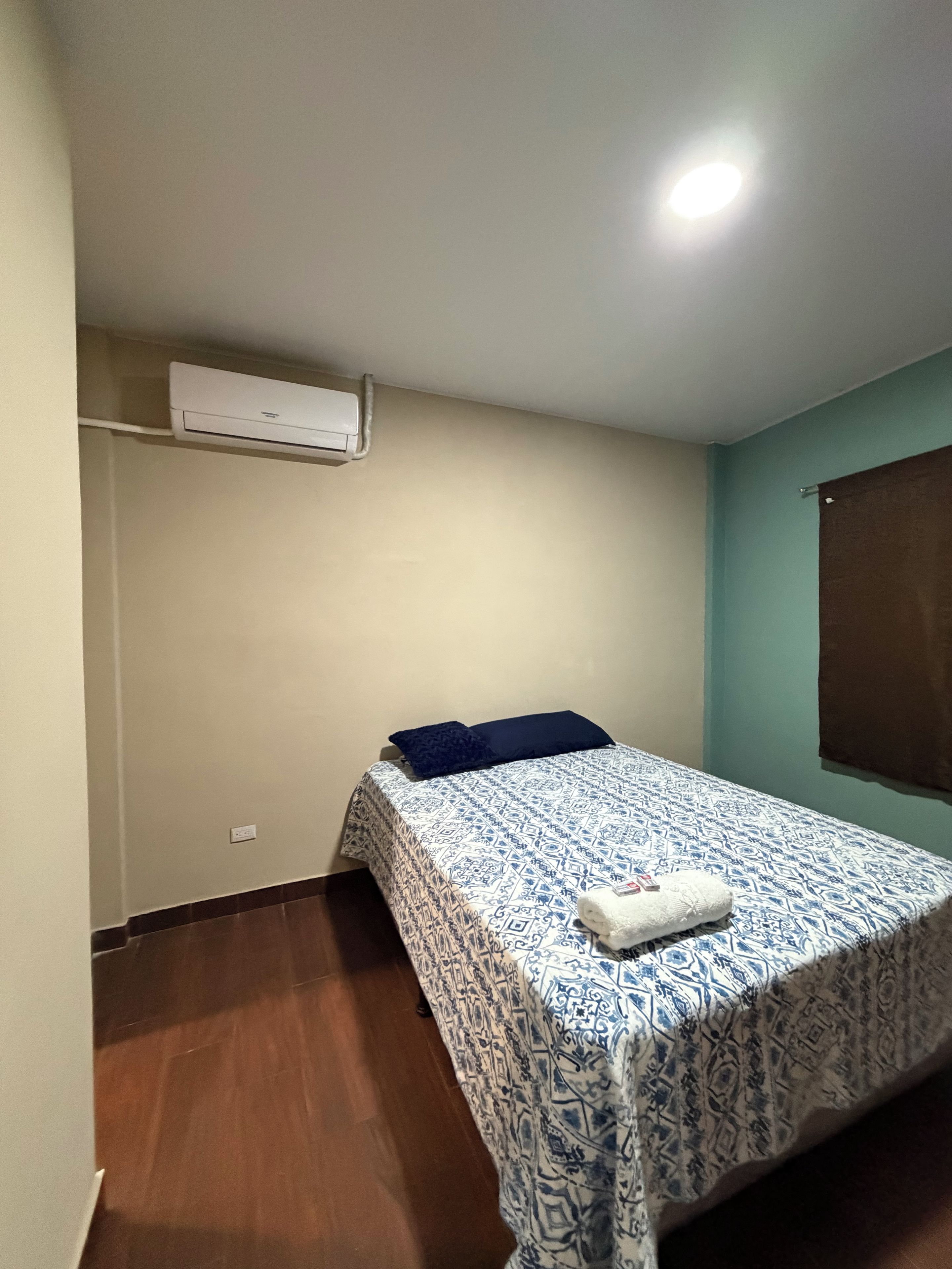 Une petite chambre avec un lit simple, une couvre-lit à motifs, un climatiseur fixé au mur et un rideau marron.