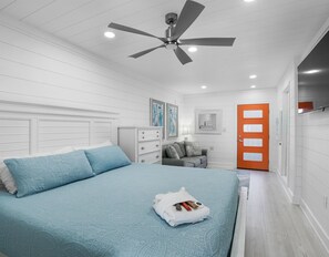 Une chambre à coucher avec un lit, un ventilateur de plafond, une commode, un canapé gris, une porte et une fenêtre.