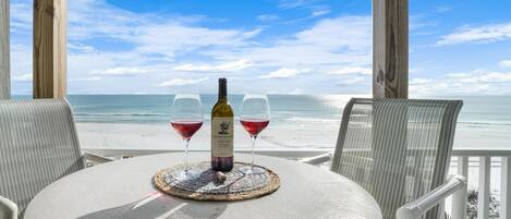 Mesa redonda con una botella de vino y dos copas sobre un tapete tejido, con vista a una playa.