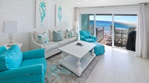 A modern living room with a white sofa, teal armchairs, and a white coffee table. There are two abstract paintings on the wall, a lamp on a side table, and a view of the ocean through a sliding glass door. - 12B - 2BR $700+ in freebies incl golf, On Beach (Miramar Beach)