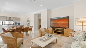 A modern living room with a dining area, a flat-screen TV, and a kitchen in the background. - Catherine Hill Bay - Ocean Views & Coastal Living (Catherine Hill Bay)
