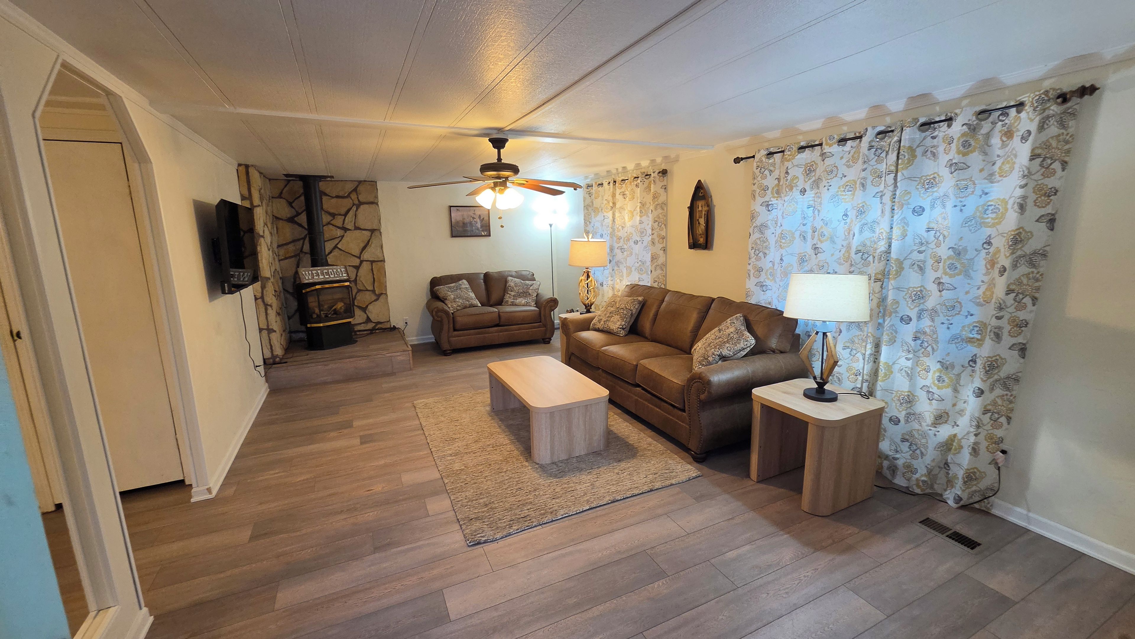 A living room with a sofa, a coffee table, a fireplace, a ceiling fan, and a patterned curtain.