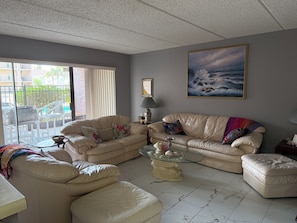 A living room with a beige sofa set, a glass coffee table, and a view of a balcony with outdoor furniture. - 2-bedroom condo in serene South Padre Island just steps from the beach. (South Padre Island)