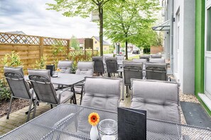 Ein Terrassenbereich im Freien mit mehreren Tischen und Stühlen, umgeben von einem Holzzaun und Grünpflanzen.