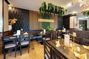 Ein Restaurantinneres mit Holztischen und -stühlen, einer grünen Wand mit hängenden Pflanzen und einem Schild mit der Aufschrift 'MISTRO'.
