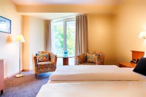 Ein Hotelzimmer mit einem Bett, zwei Sesseln, einem kleinen Tisch, einer Lampe und einem Fenster mit Vorhängen.