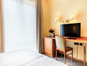Ein Hotelzimmer mit Bett, Schreibtisch mit Fernseher, Stuhl, kleiner Schrank und einem Fenster mit Vorhängen.
