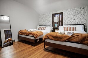 Quarto com duas camas, cada uma com um cobertor castanho e roupa de cama branca. Há um espelho na parede à esquerda e uma porta que dá para outro quarto.