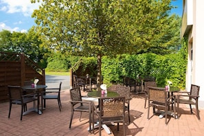 Ein Terrassenbereich im Freien mit Korbmöbeln und einem kleinen Tisch, umgeben von Grünpflanzen und einem Holzzaun.
