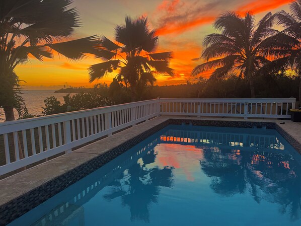 A swimming pool with a white deck and a view of the sunset over the ocean.