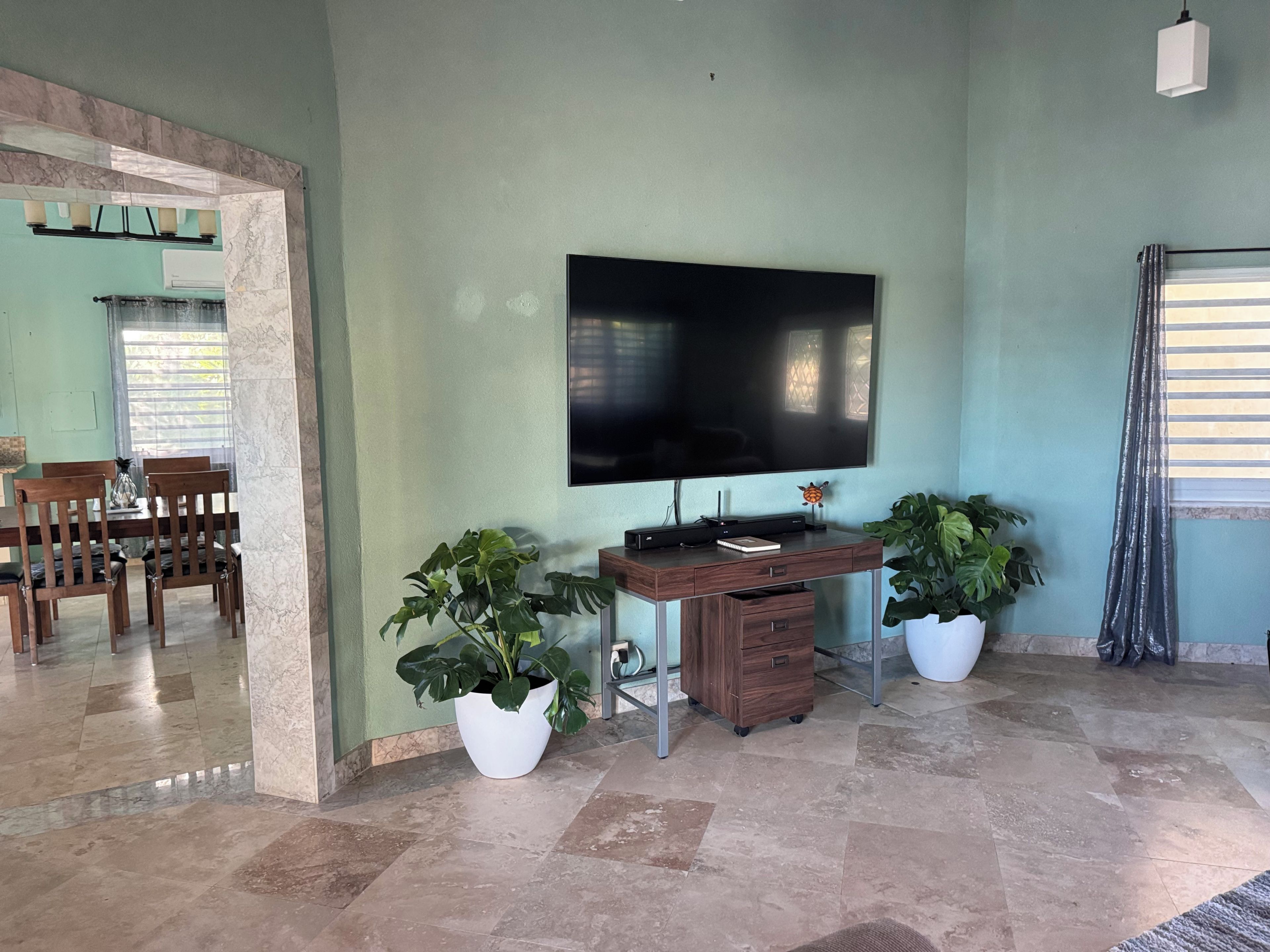 A living room with a flat-screen TV, wooden furniture, potted plants, and a tiled floor.