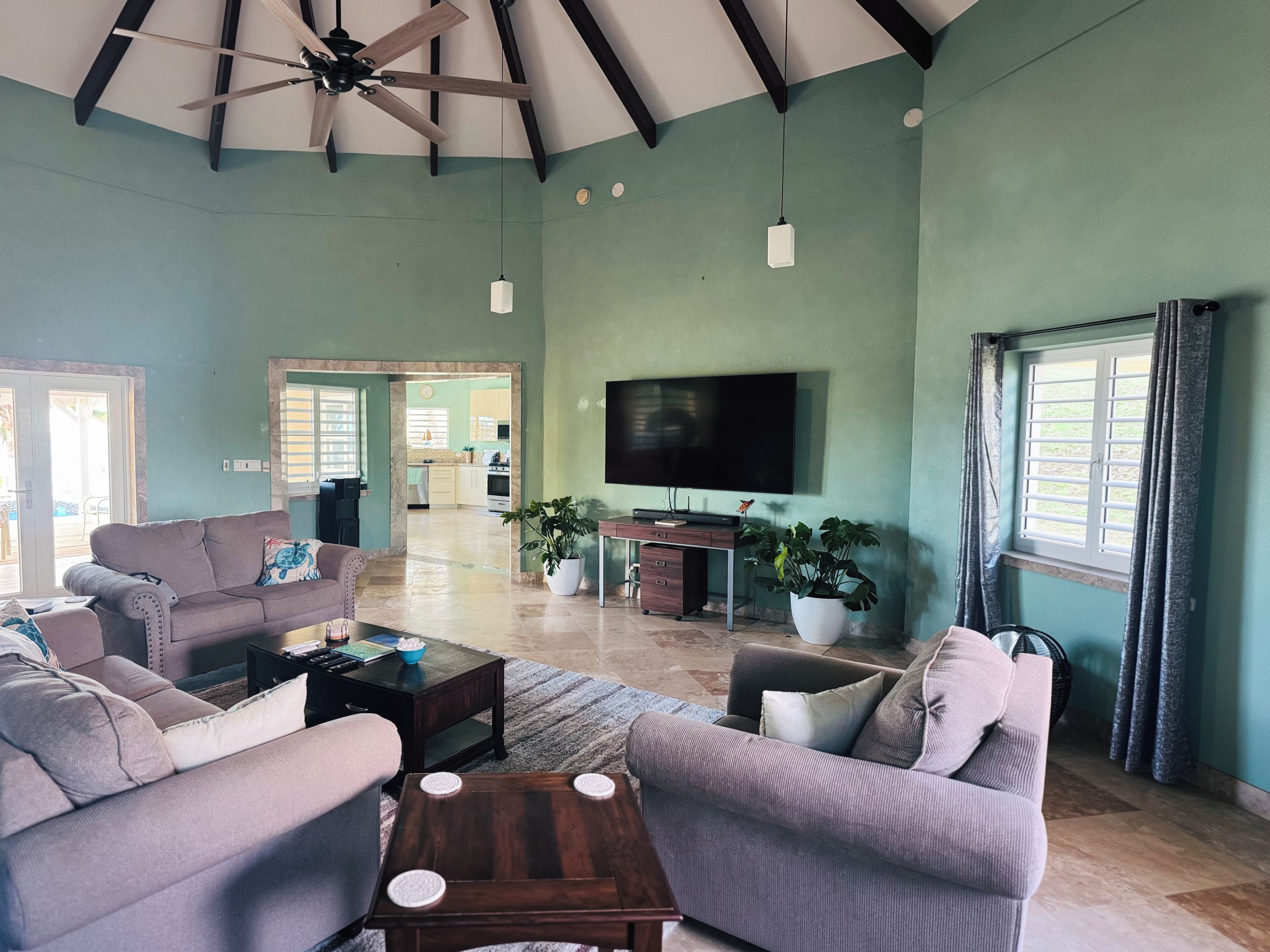A spacious living room with a high ceiling, a flat-screen TV mounted on the wall, and a large window with curtains.